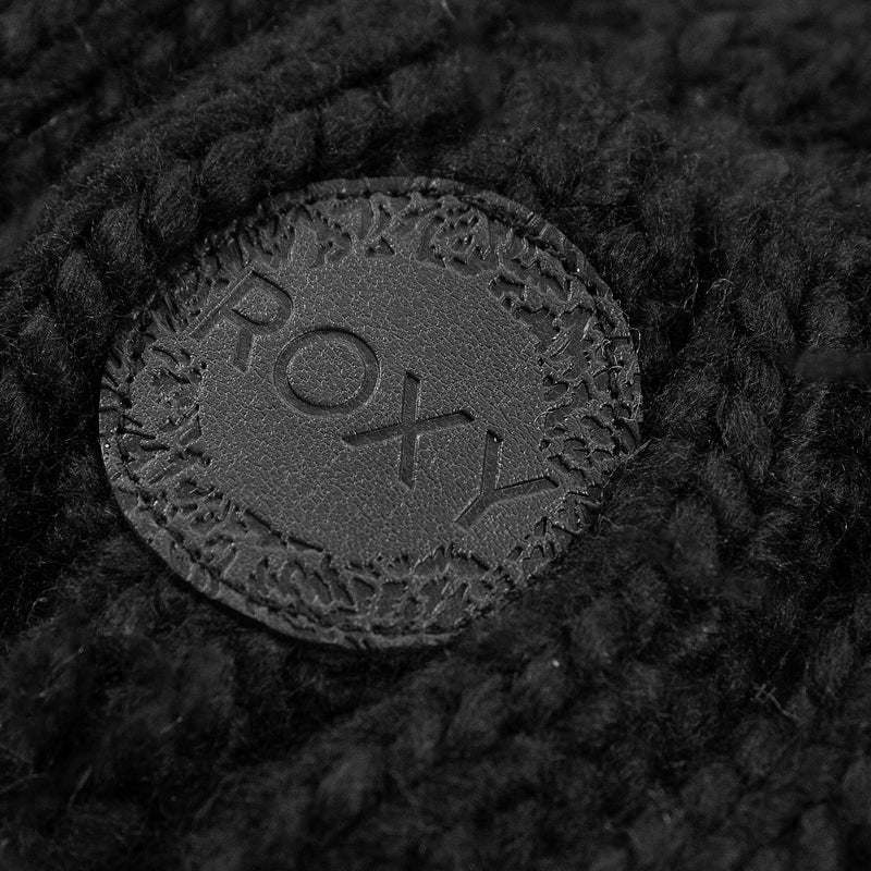 Close-up of a black circular leather patch with the brand name “Roxy” embossed on it, attached to the thick cable knit fabric of the Tram Beanie.
