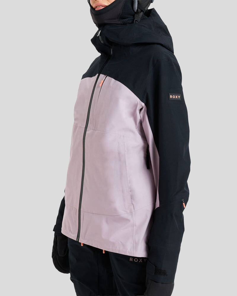 A person wearing the Roxy Gore-Tex Lunalite Jacket, a two-tone snowboard jacket in light pink and black with a hood, gloves, and high collar. Designed for mountain performance, this waterproof and breathable jacket displays the ROXY logo on the sleeve and near the hem. The background is plain light gray.