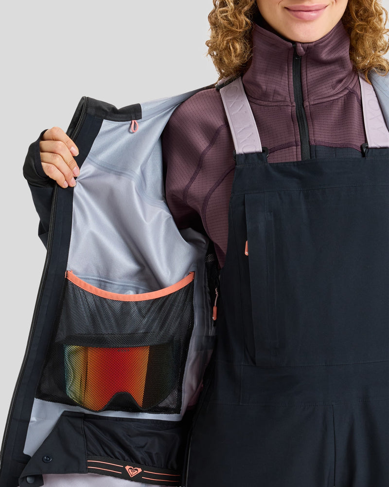 A person opens a black Roxy Gore-Tex Lunalite Jacket, revealing the interior mesh pocket holding a pair of ski goggles with an orange lens. Only the lower face and torso are visible, emphasizing this waterproof, breathable jacket engineered by Roxy for peak mountain performance.