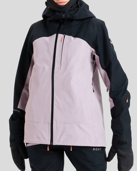 A person wearing a light purple and black Roxy Gore-Tex Lunalite Jacket, crafted from recycled polyester, along with black gloves, stands against a plain light gray background. This waterproof and breathable jacket features a hood and several zippered pockets.