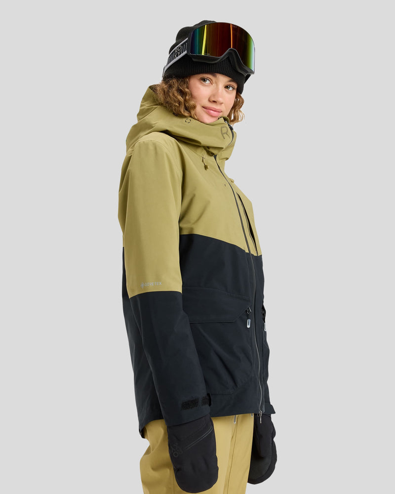 Wearing the Roxy Gore-Tex Stretch Purelines Jacket, a person outfitted with ski goggles, tan pants, gloves, and a hood stands against a plain gray background, smiling and gazing slightly to the side.