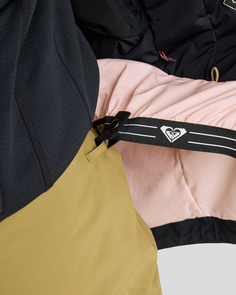 Close-up of the interior of the Roxy Gore-Tex Stretch Purelines Jacket, highlighting its pink lining, black waistband with a white Roxy logo, and part of tan pants. The waistband features both a zipper and a snap button.