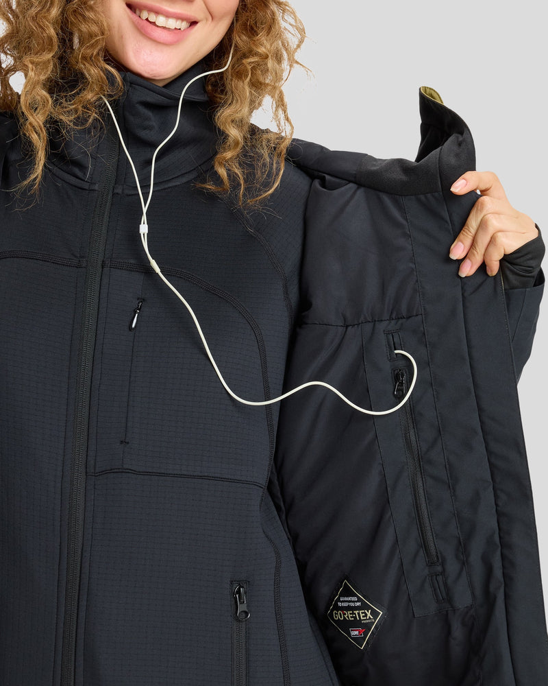 A person smiles while holding open a black Roxy Gore-Tex Stretch Purelines Jacket, revealing the interior pocket with a white earbud cord running from inside the jacket to their ear.
