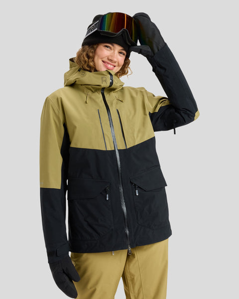 A person in a green and black Roxy Gore-Tex Stretch Purelines Jacket and matching pants smiles while lifting their ski goggles. They wear black gloves and a beanie, standing against a plain light gray background.