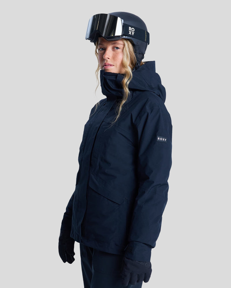 A person wearing a black Roxy Gore-Tex Stormday Jacket with PrimaLoft insulation, black gloves, and a black ski helmet with goggles stands against a plain light background, looking slightly to the side.