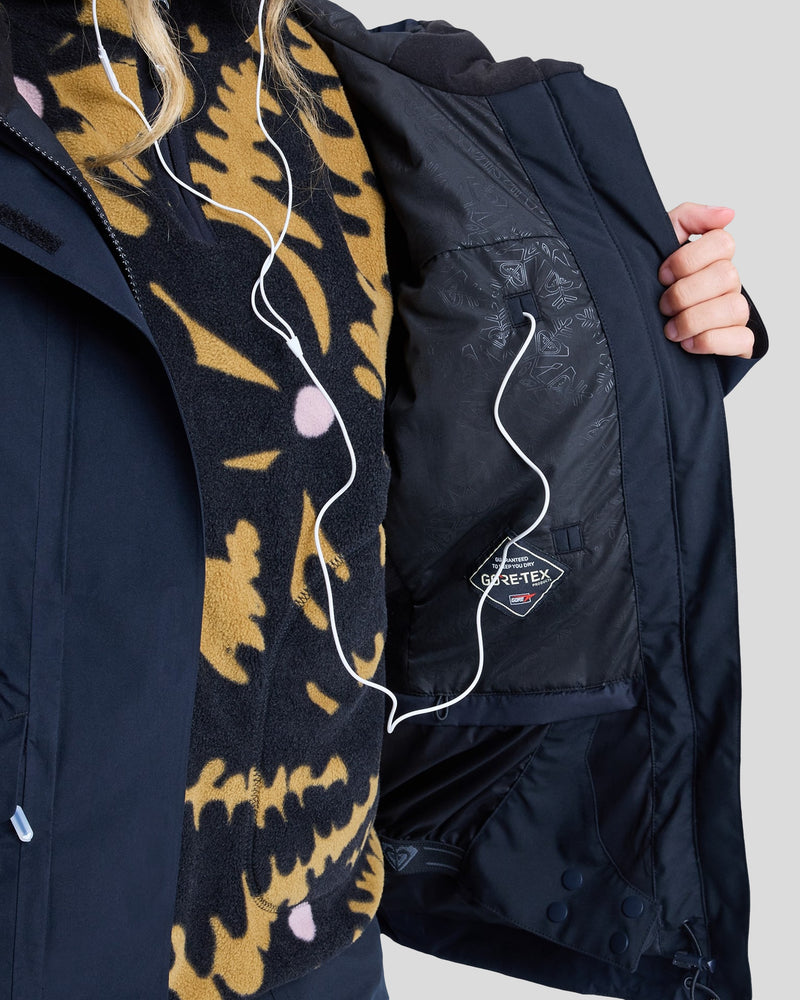 A person wearing a black and yellow patterned fleece and the Roxy Gore-Tex Stormday Jacket holds open the jacket to reveal its inner GORE-TEX lining, with white earphones hanging from their neck.