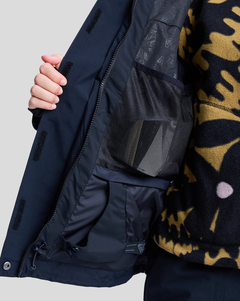 A person opens a Roxy Gore-Tex Stormday Jacket in navy blue, revealing mesh and fabric inner pockets, and wears a brown and black patterned fleece sweater underneath.