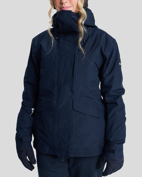 A person wearing the Roxy Gore-Tex Stormday Jacket in dark blue, featuring PrimaLoft insulation, multiple pockets, a high collar, and a hood, along with gloves, stands against a plain light gray background. Their face is partially visible beneath wavy blond hair.