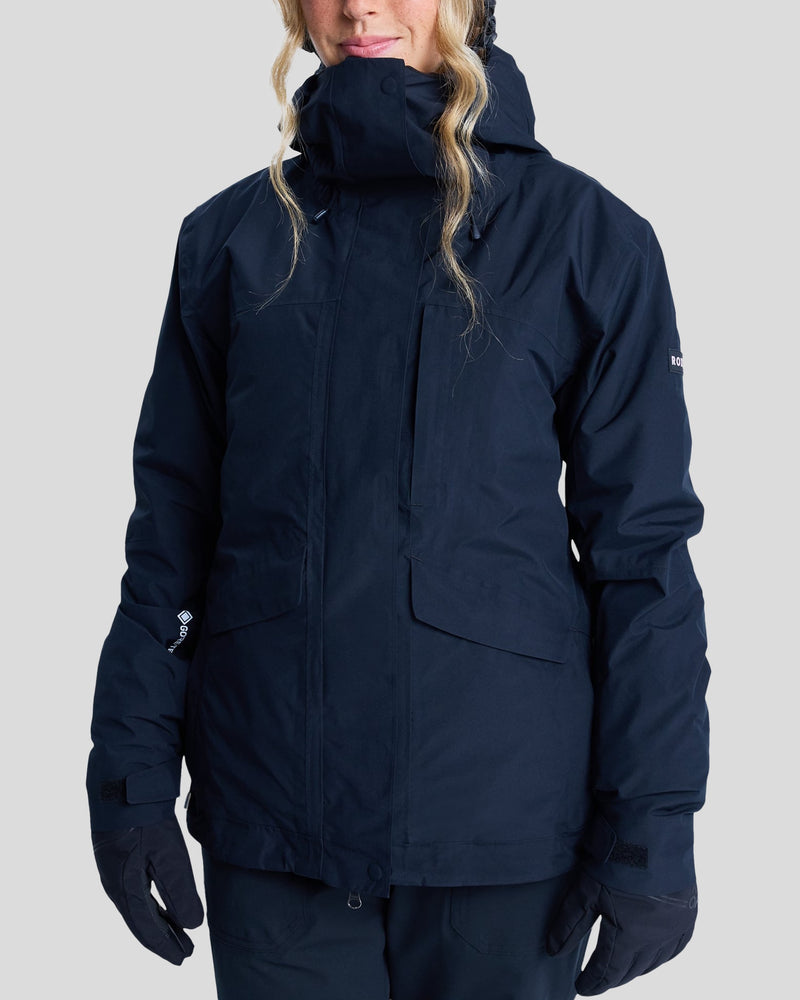A person wearing the Roxy Gore-Tex Stormday Jacket in dark blue, featuring PrimaLoft insulation, multiple pockets, a high collar, and a hood, along with gloves, stands against a plain light gray background. Their face is partially visible beneath wavy blond hair.