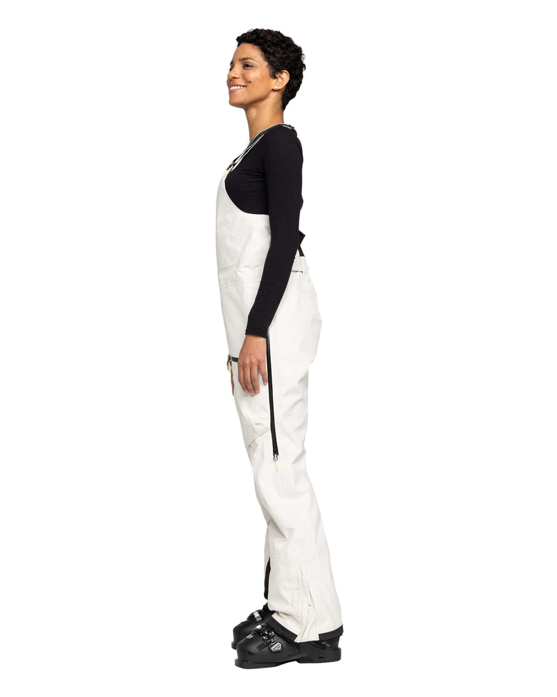 A person stands in profile, smiling, wearing a black long-sleeve top, black boots, and the Roxy Gore-Tex 3L Lunalite Bib—white women’s snow bib with side zippers—against a plain white background.