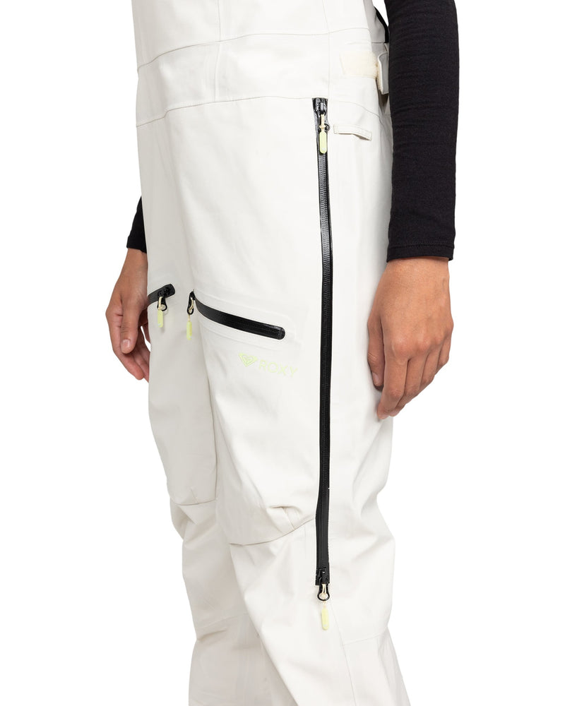 A person is wearing the ROXY Gore-Tex 3L Lunalite Bib in white, featuring black zippers, paired with a black long-sleeve shirt. Only the lower torso and legs are visible. This waterproof women’s snow bib from ROXY offers reliable protection in wintry conditions.