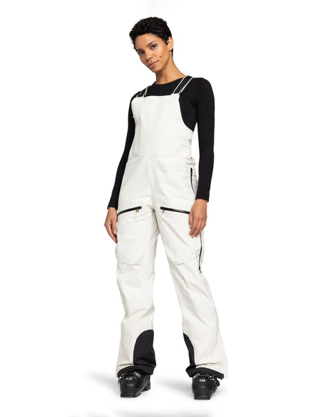 A person stands facing forward against a plain white background, wearing the Roxy Gore-Tex 3L Lunalite Bib—white women’s snow bib overalls crafted from waterproof, breathable fabric—layered over a black long-sleeve shirt and paired with black boots.