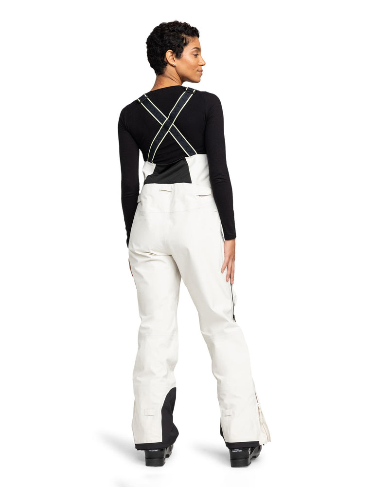 A person stands facing away, wearing a black long-sleeve top and the Roxy Gore-Tex 3L Lunalite Bib—white women’s snow pants with black suspenders crossing in the back. The bib is crafted from waterproof GORE-TEX 3L fabric, and they are also wearing black snow boots.