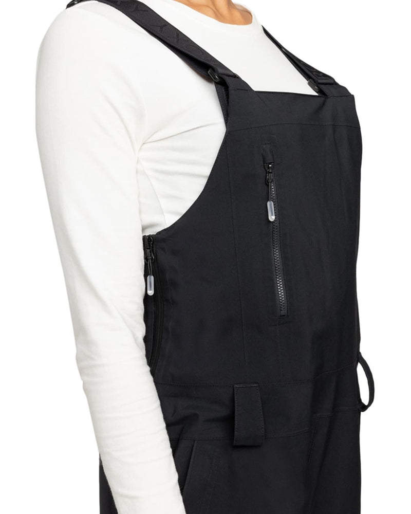 A person is shown from the shoulders to the hips against a white background, wearing a long-sleeve white shirt paired with the Roxy Gore-Tex Stretch Prism Bib—black overalls featuring zipper pockets and crafted from waterproof, breathable fabric.
