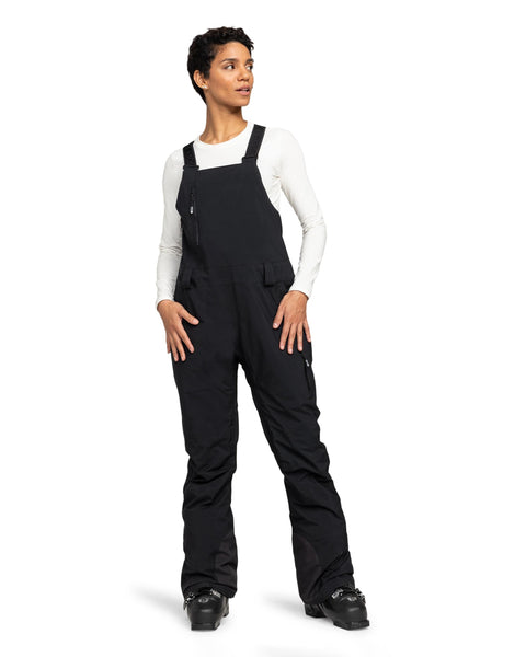 A person wearing a white long-sleeve shirt, black Roxy Gore-Tex Stretch Prism Bib ski overalls with waterproof breathable protection, and black ski boots stands against a plain white background, looking slightly to the left.