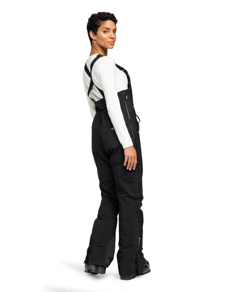 A person stands facing backward, looking over their shoulder while wearing Roxy’s Gore-Tex Stretch Prism Bib in black and a white long-sleeve shirt. The outfit is completed with black boots, and the waterproof, breathable GORE-TEX fabric delivers comfort and protection in any weather. The background is plain white.
