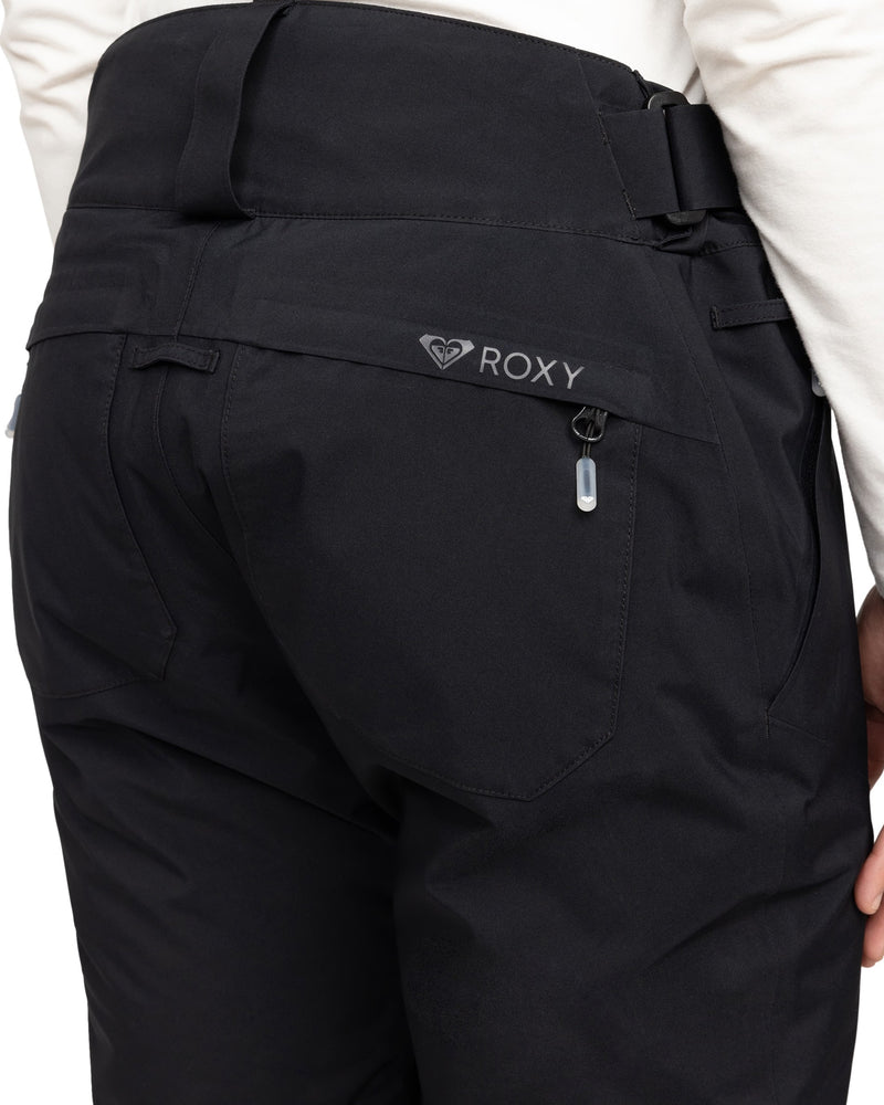 Close-up of a person wearing black Roxy Gore-Tex Spridle Pants, highlighting the zipped back pocket with the Roxy logo above it. The person is also dressed in a white top.