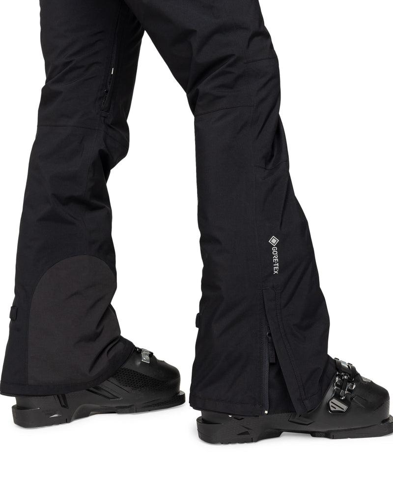 A person wearing black Roxy Gore-Tex Spridle Pants with black ski boots, standing on a white background. Only the lower legs and feet are visible, highlighting the waterproof and breathable features of these snow pants.