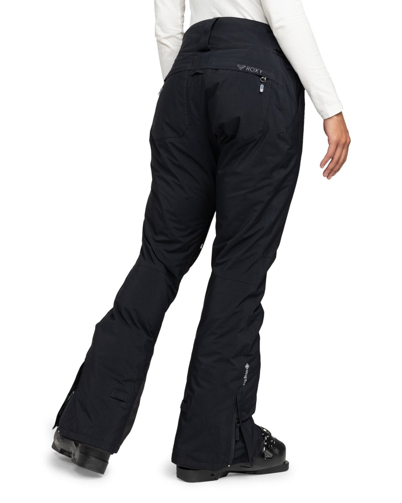 A person is shown from the back wearing Roxy Gore-Tex Spridle Pants in black, paired with black boots. The pants feature a slightly flared bottom and zippered pockets. The individual is also dressed in a white long-sleeve shirt.