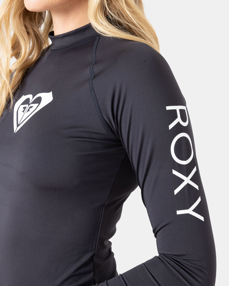 A person with long blonde hair wears the Roxy Whole Hearted LS Rashguard, made from recycled stretch fabric, featuring the Roxy logo on the chest and "ROXY" in white on the sleeve.