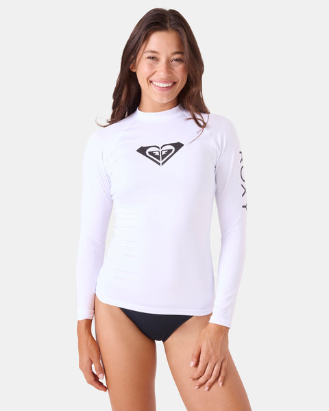 A woman smiles against a plain background wearing the Roxy Whole Hearted LS Rashguard, featuring a fitted silhouette, long sleeves, black logo on the chest, ROXY print on the sleeve, and paired with black swim bottoms.