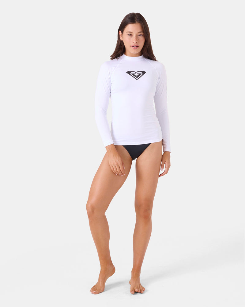 A woman stands barefoot against a white background, wearing the Roxy Whole Hearted LS Rashguard with a fitted silhouette and black chest logo, paired with black bikini bottoms.