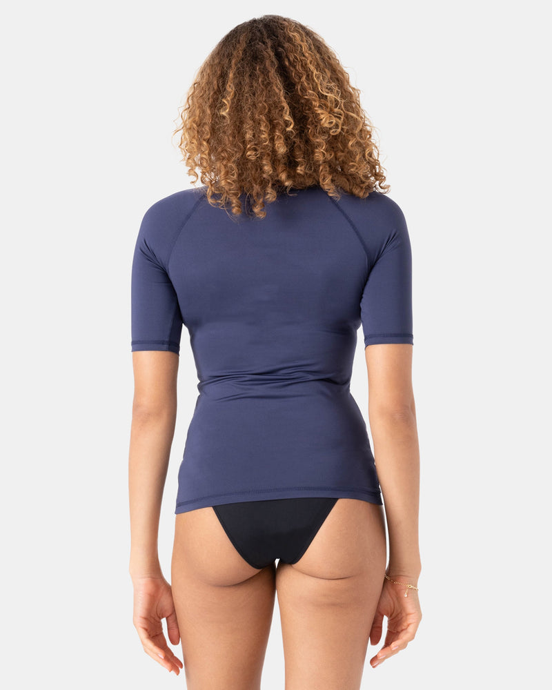 A person with curly hair is seen from the back, wearing the Roxy Whole Hearted Ss Rashguard in navy and black bikini bottoms, made from recycled stretch fabric, set against a plain white background.