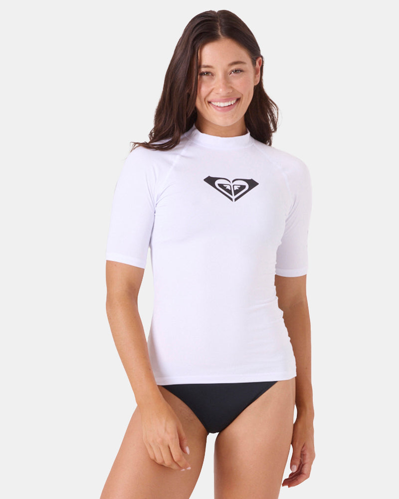 A woman smiles in a fitted Roxy Whole Hearted Ss Rashguard, crafted from recycled polyester, paired with black bikini bottoms against a plain light background.