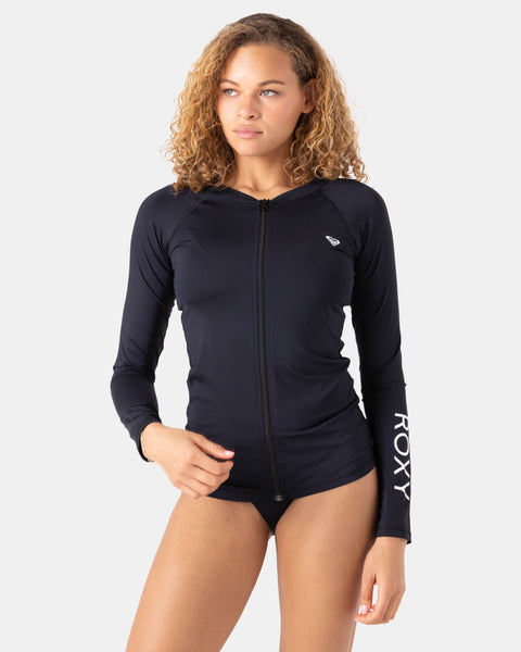 A woman with curly hair wears the Roxy New Essentials Ls Zip Rashguard, a black long sleeve style with ROXY branding on one sleeve, standing against a plain light background.