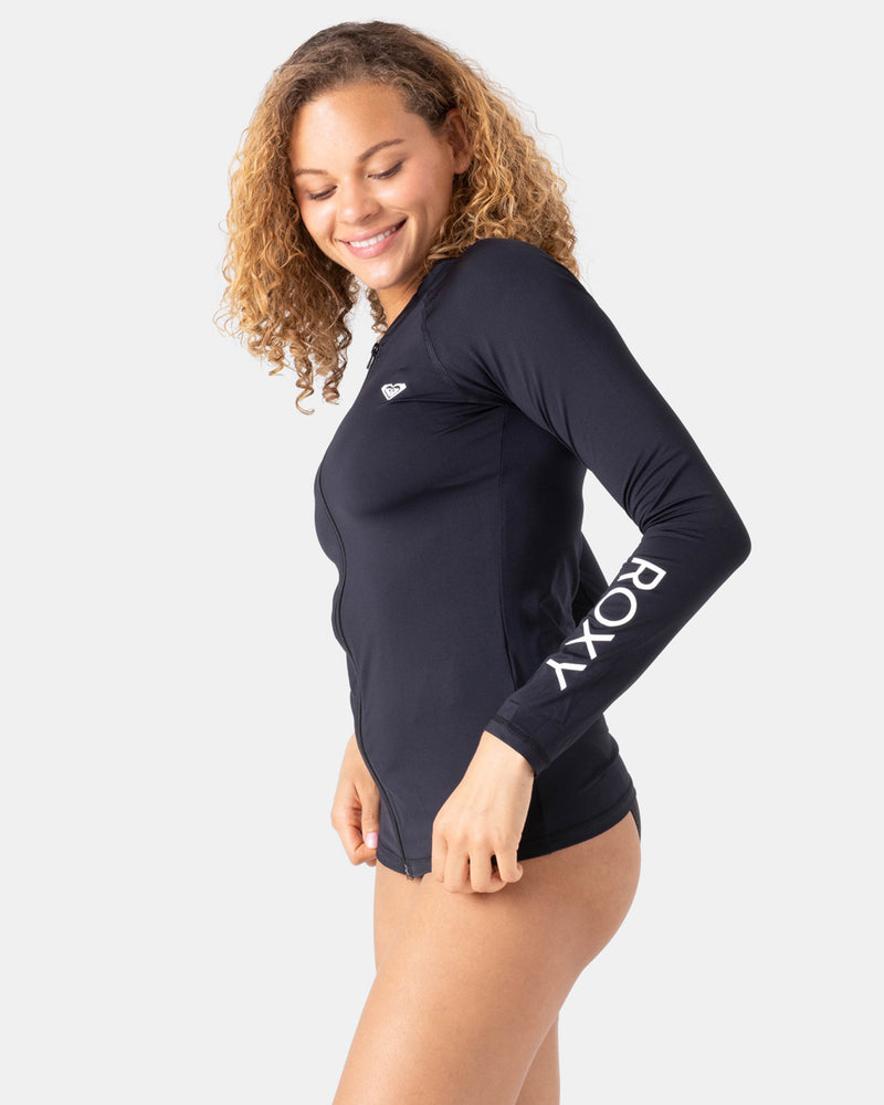 A woman in a Roxy New Essentials LS Zip Rashguard with ROXY on the sleeve smiles and looks down, standing sideways to show off the sleek profile of the recycled-stretch swimsuit against a plain light background.