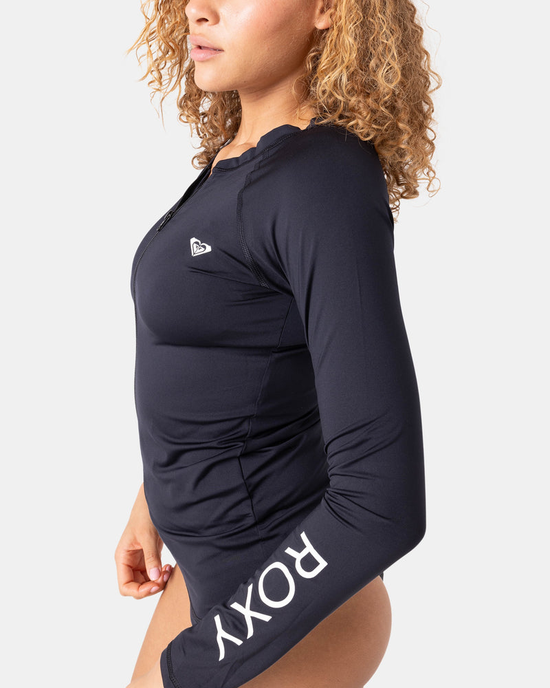 A person with curly hair wears the black Roxy New Essentials LS Zip Rashguard with long sleeves and a small chest logo, featuring ROXY on the sleeve, made from recycled-stretch fabric, standing against a plain white background.