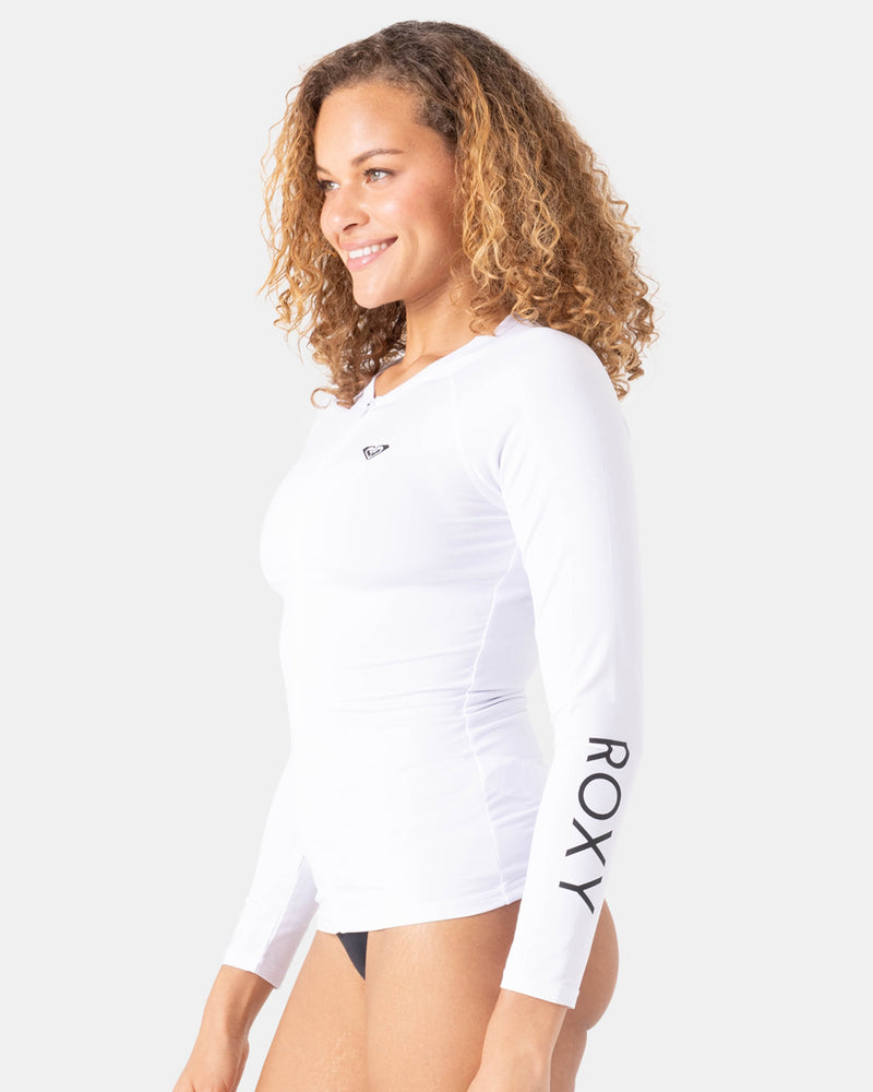 A woman with curly hair wears a fitted Roxy New Essentials LS Zip Rashguard in bright white, featuring "ROXY" on the left sleeve and a chest logo. She smiles and faces sideways against a plain background.