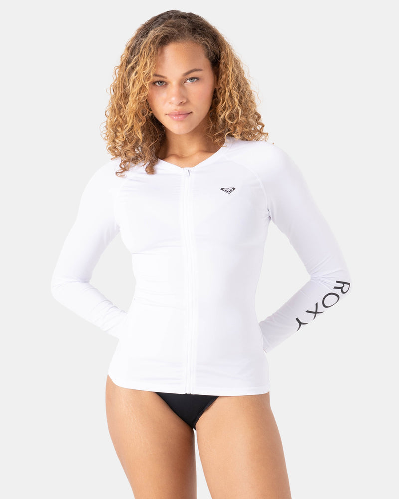 A woman with curly hair wears a fitted New Essentials Ls Zip Rashguard by Roxy in Bright White and black bikini bottoms, standing against a plain light gray background.