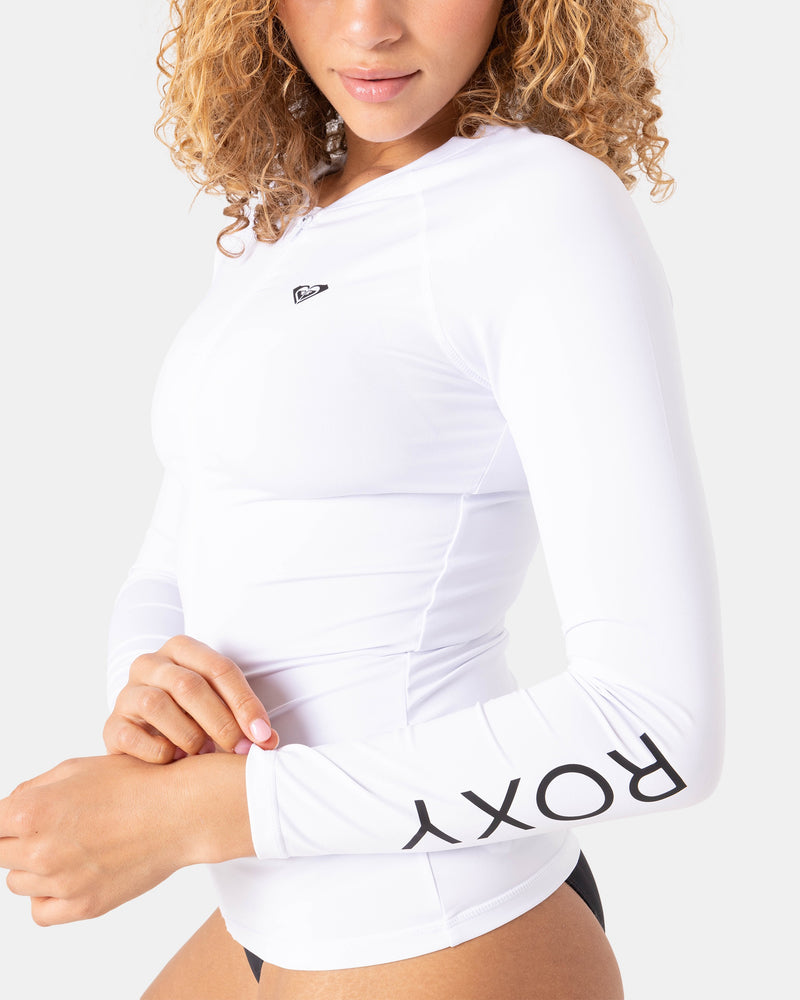 A person with curly hair wears a fitted, long-sleeve New Essentials LS Zip Rashguard by Roxy in bright white, featuring "ROXY" printed in black on the sleeve and a small chest logo, paired with black swim bottoms.