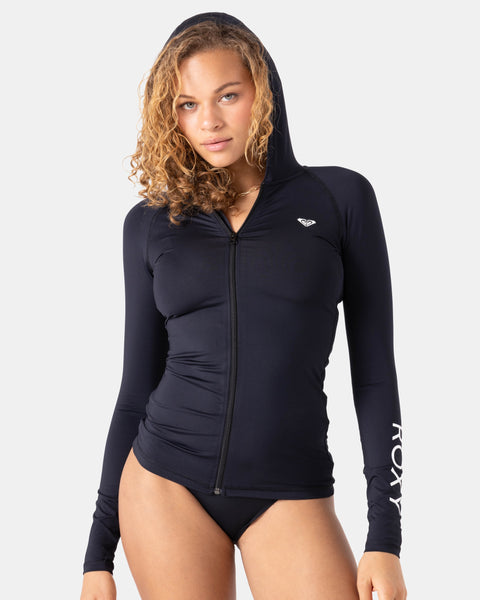 A woman with curly hair wears the Roxy New Essentials Ls Zip Hooded rash guard, featuring ROXY on the left sleeve and a chest logo, paired with black bikini bottoms against a plain light background.