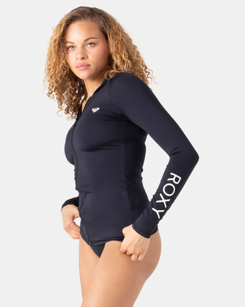 A woman with curly hair wears a Roxy New Essentials Ls Zip Hooded cover-up with ROXY printed on the sleeve and a small logo on the chest, standing against a plain white background.