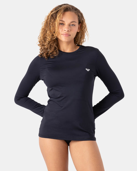A person with curly hair stands against a plain background, wearing the Roxy New Enjoy Waves Ls fitted long-sleeve black rashguard made from recycled polyester and black bikini bottoms, hands on hips with a neutral expression.