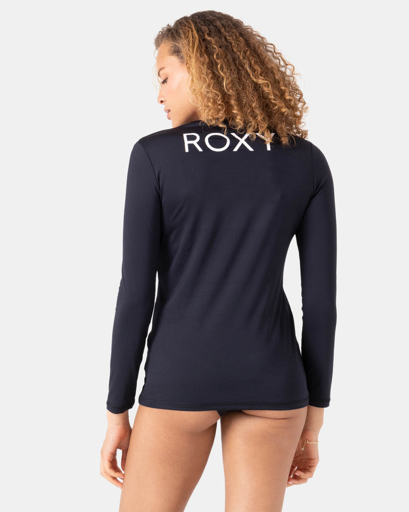 A woman with curly hair is shown from the back wearing the Roxy New Enjoy Waves Ls long-sleeve black rashguard made from recycled polyester, featuring "ROXY" in white letters across the upper back, against a plain white background.