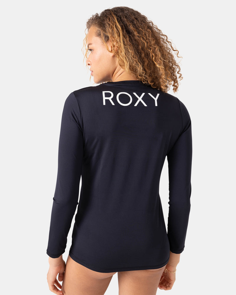 A person with curly hair wears the Roxy New Enjoy Waves Ls, a long-sleeve black rashguard made from recycled polyester with ROXY printed in large white letters on the back, standing against a plain white background.