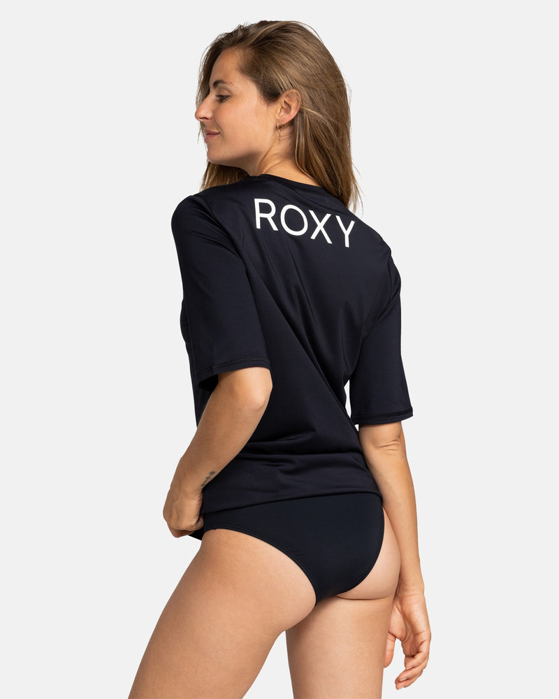 A woman in profile smiles, wearing the Roxy New Enjoy Waves Ss black surf rashguard with ROXY printed on the back and matching black bikini bottoms, set against a plain light background.