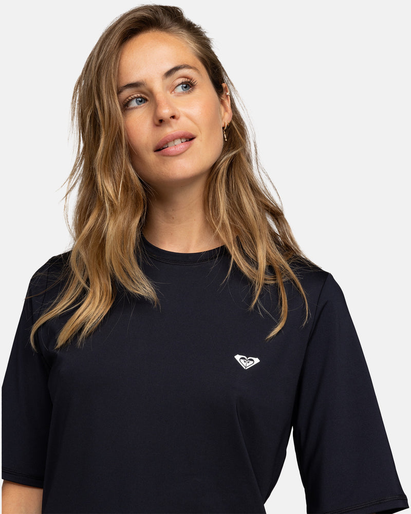 A woman with long, wavy blond hair wears the black Roxy New Enjoy Waves Ss rashguard, featuring a small white heart logo on the chest, looking up and to her left against a plain background.