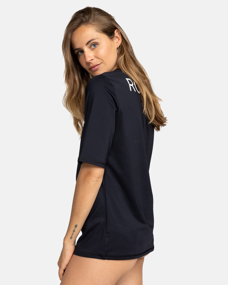 A woman with long light brown hair models the Roxy New Enjoy Waves Ss black recycled rashguard, glancing over her shoulder at the camera. The back features white 'RU' text, and she stands against a plain light background.