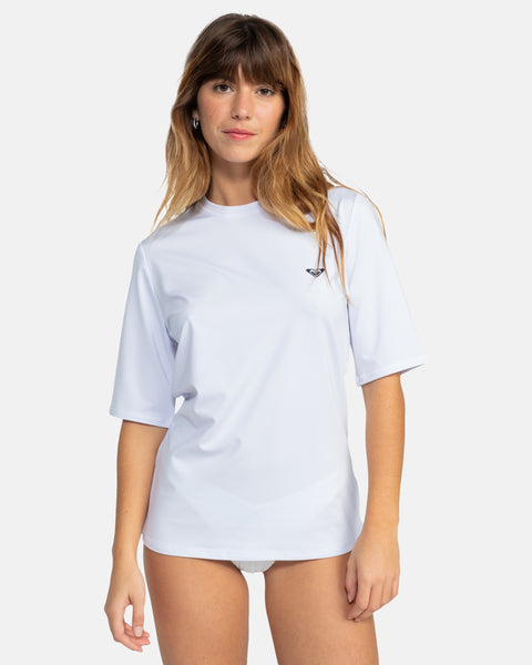 A woman with long brown hair models the Roxy New Enjoy Waves Ss, a loose-fitting white short-sleeve rashguard with a small chest logo, standing against a plain white background.