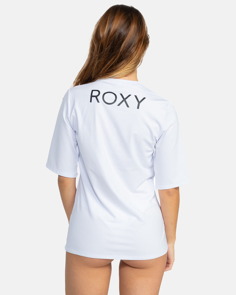 A woman with long brown hair stands with her back to the camera, wearing the Roxy New Enjoy Waves Ss—a loose, short-sleeve white T-shirt with a bold black ROXY logo on the upper back, styled in classic surf fashion.