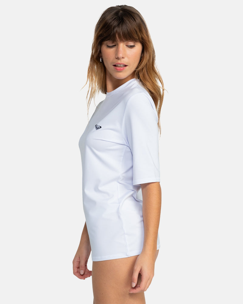 A woman with long brown hair models the Roxy New Enjoy Waves Ss light blue short-sleeve rashguard, standing sideways against a plain white background.