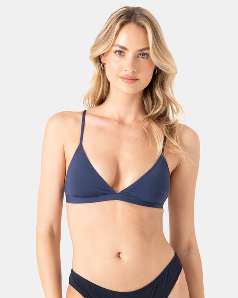 A woman with long, wavy blonde hair models the Roxy Sd Beach Classics Fixed Tri navy blue triangle bikini, made from recycled nylon. She stands against a light grey background, gazing at the camera with a neutral expression.