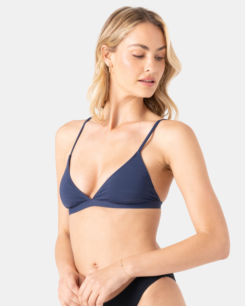A woman with blonde hair models the Roxy Sd Beach Classics Fixed Tri navy triangle bikini top with black bottoms, made from recycled nylon, looking down and slightly to the side against a plain light background—perfect for swim days.