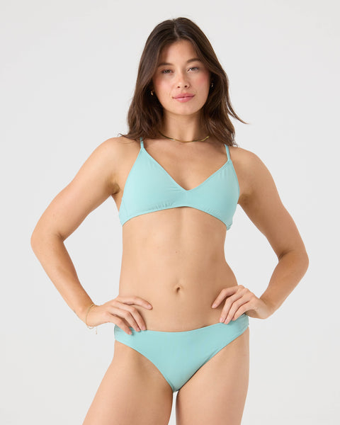 A woman with long brown hair and a neutral expression poses against a plain light background, wearing the Roxy SD Beach Classics Short Sleeve Athletic Triangle Bikini Top in light blue recycled nylon.