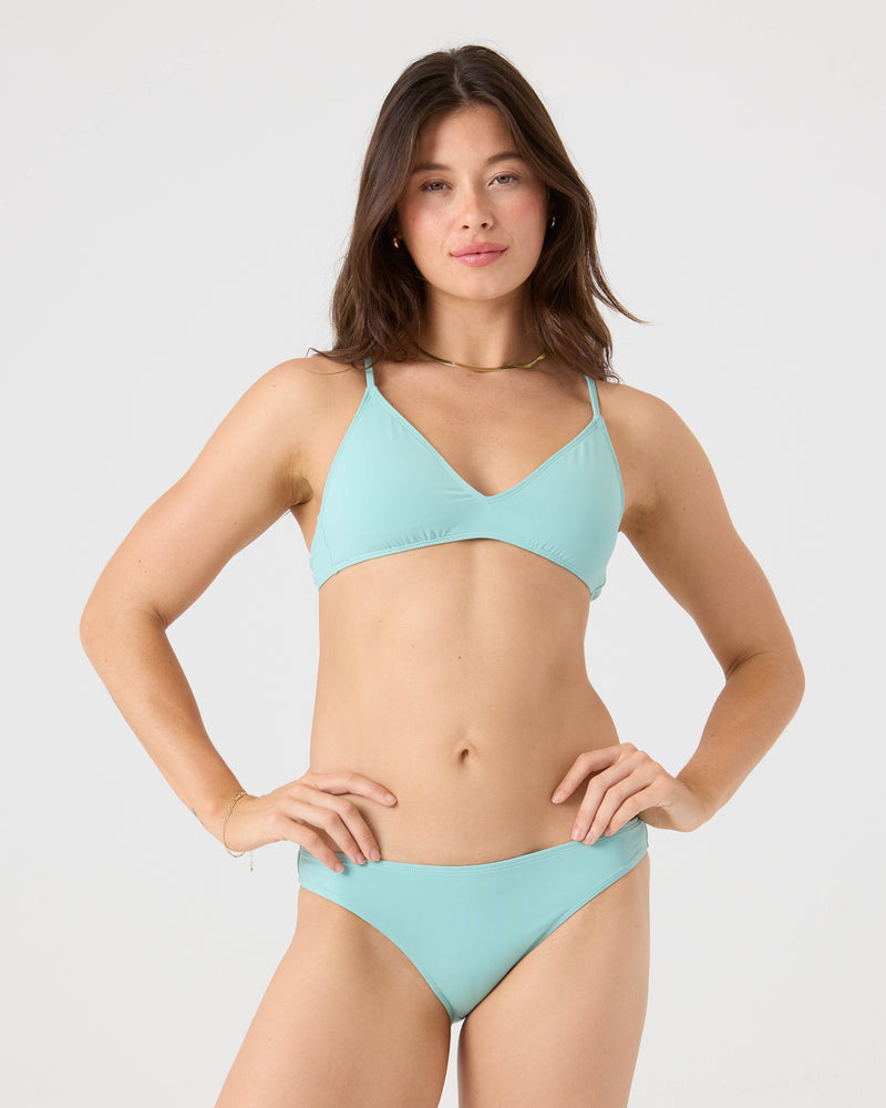 A woman with long brown hair and a neutral expression poses against a plain light background, wearing the Roxy SD Beach Classics Short Sleeve Athletic Triangle Bikini Top in light blue recycled nylon.