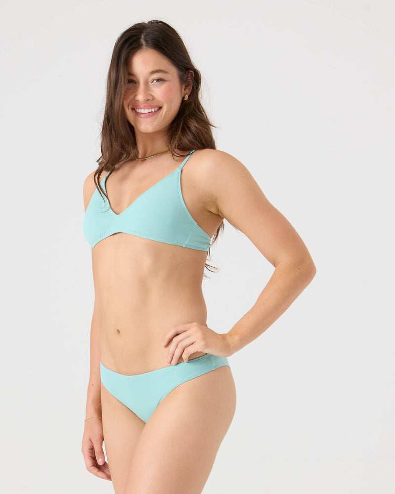 A woman with long brown hair smiles while posing in a light blue Roxy SD Beach Classics Short Sleeve Athletic Triangle Bikini Top against a plain white background, one hand on her hip and standing slightly angled to the side.